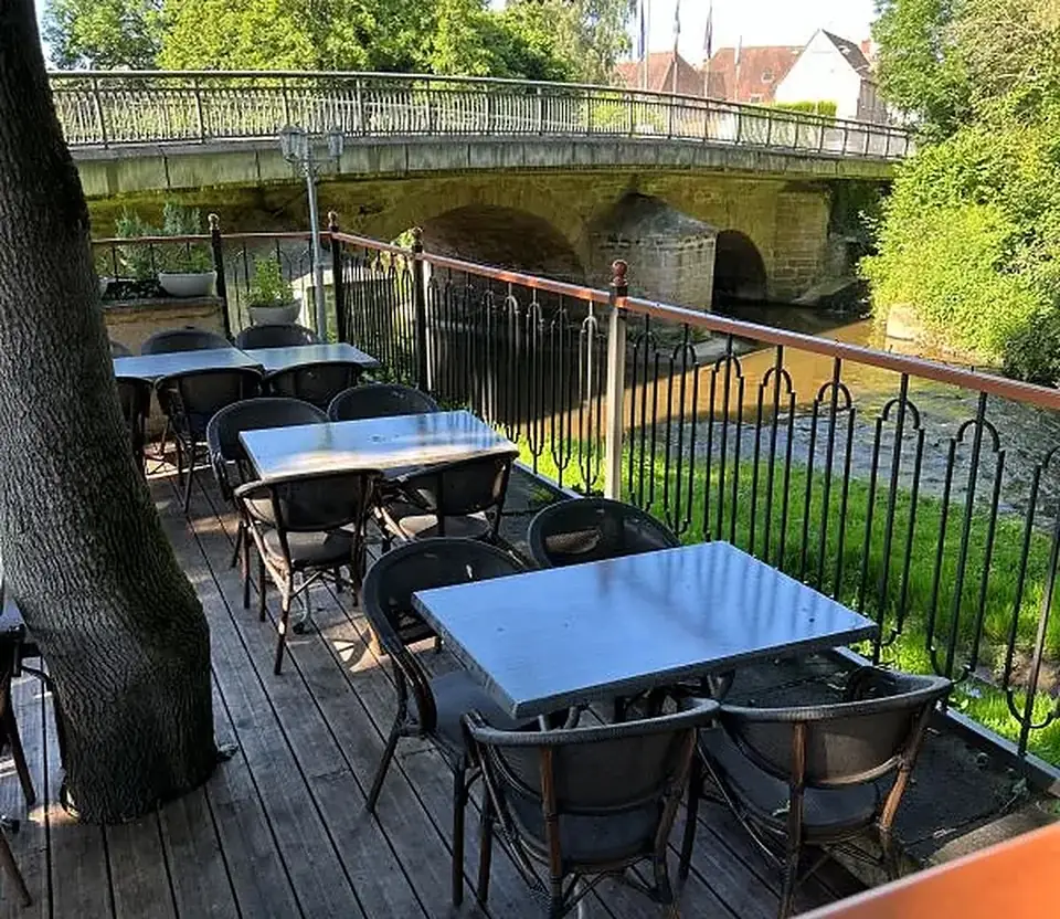 La terrasse du restaurant : des tables et des chaises sur un plancher de bois, au pied d’un grand arbre, avec la rivière et un pont de pierre derrière